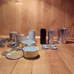 Różne rodzaje metalowych puszek i pokrywek na drewnianym tle, widoczne puszki aluminiowe, stalowe i czarne plastikowe, niektóre z pokrywkami typu easy-open