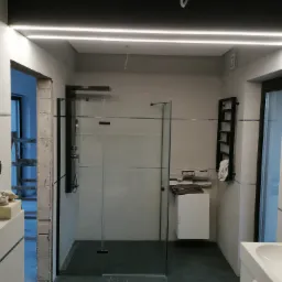 Nowoczesna łazienka w trakcie remontu z kabiną prysznicową typu walk-in, białą armaturą i grzejnikiem drabinkowym; widoczne niedokończone obramowanie drzwi i przewody elektryczne.