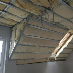 Izolacja poddasza wełną mineralną, widoczne metalowe profile konstrukcyjne i okno dachowe w trakcie montażu, oświetlenie dzienne.