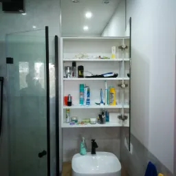 Nowoczesna łazienka z białą umywalką nablatową, drewnianym blatem i szafką z lustrem w zabudowie, częściowo wypełnioną kosmetykami i akcesoriami, obok kabina prysznicowa ze szklanymi drzwiami.