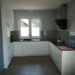 Nowoczesna, minimalistyczna kuchnia z białymi frontami szafek, czarnym blatem i okapem, widok na zewnątrz przez okno nad zlewem. Na blacie stoją butelki wody i czajnik.