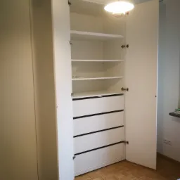 Otwarta biała szafa z półkami i szufladami z czarnymi uchwytami, widoczna na tle parkietu i białej ściany, oświetlona wiszącą lampą.