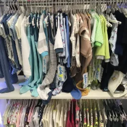 Regał sklepowy z wieszakami, na których wisi różnorodna dziecięca odzież niemowlęca w pastelowych kolorach, widoczne metki z cenami.