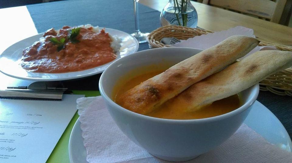 Zupa krem w białej miseczce z dwoma grzankami, na stole obok danie z sosem i ryżem, kieliszek i menu restauracji.