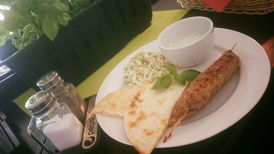 Porcja grillowanego mięsa na szpadce, podana z plackami pita, surówką i sosem w białej miseczce, udekorowana listkiem bazylii, obok solniczka i pieprzniczka z logo Pepsi.