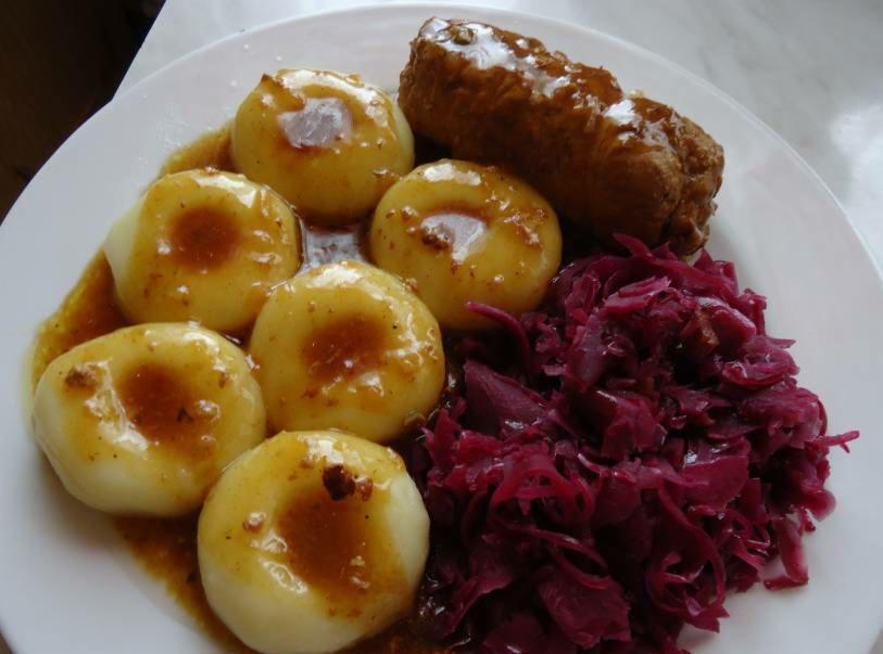 Talerz z kluskami śląskimi polanymi sosem, pieczenią i czerwoną kapustą, ujęcie z góry, światło dzienne.