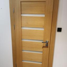 STOLARSTWO ANDREAS KAMPA - Dębowa okleina na drzwiach z poziomymi przeszkleniami, widoczna klamka i zawiasy w kolorze srebrnym, na ścianie czarny włącznik światła.