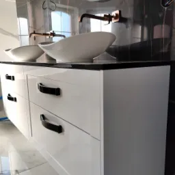 Łazienka z białymi szafkami z czarnymi uchwytami, umywalkami nablatowymi o owalnym kształcie i miedzianą armaturą na tle marmurowej ściany z okrągłym lustrem z podświetleniem.