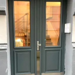 Szare dwuskrzydłowe drzwi wejściowe z szybami, widok na schody i wnętrze klatki schodowej, zamek i domofon po prawej stronie.