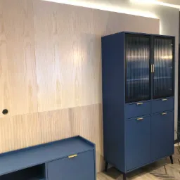 Niebieska szafka z ryflowanymi szklanymi drzwiczkami i komoda RTV z minimalistycznymi złotymi uchwytami na tle drewnianej ściany z oświetleniem LED.