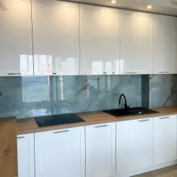 Nowoczesna kuchnia z białymi, lakierowanymi frontami szafek, drewnianym blatem i szklanym panelem ściennym imitującym marmur w odcieniach szarości i brązu, czarny zlew i bateria.