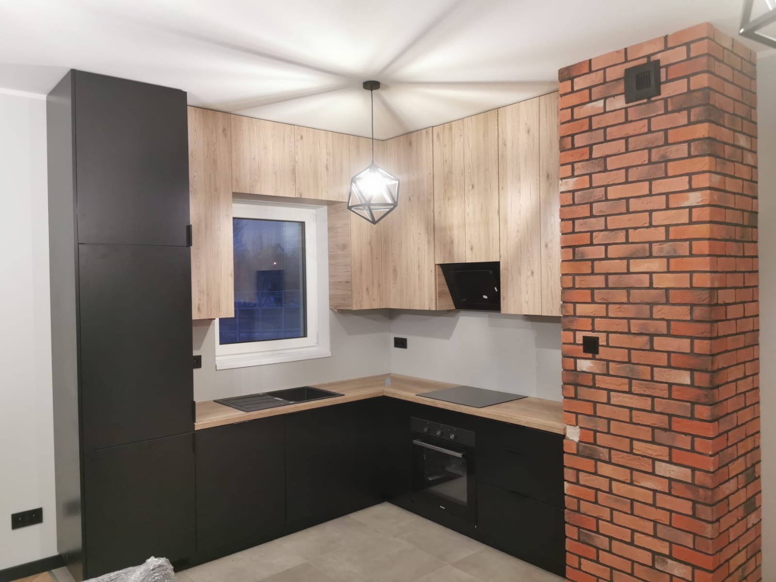 Nowoczesna kuchnia z czarnymi szafkami dolnymi i drewnopodobnymi górnymi, ceglana kolumna, minimalistyczna lampa wisząca, widok na okno.