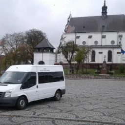 Biały bus transportowy Ford Transit na brukowanym placu, w tle biały kościół z wieżą i budynki miejskie w pochmurny dzień.