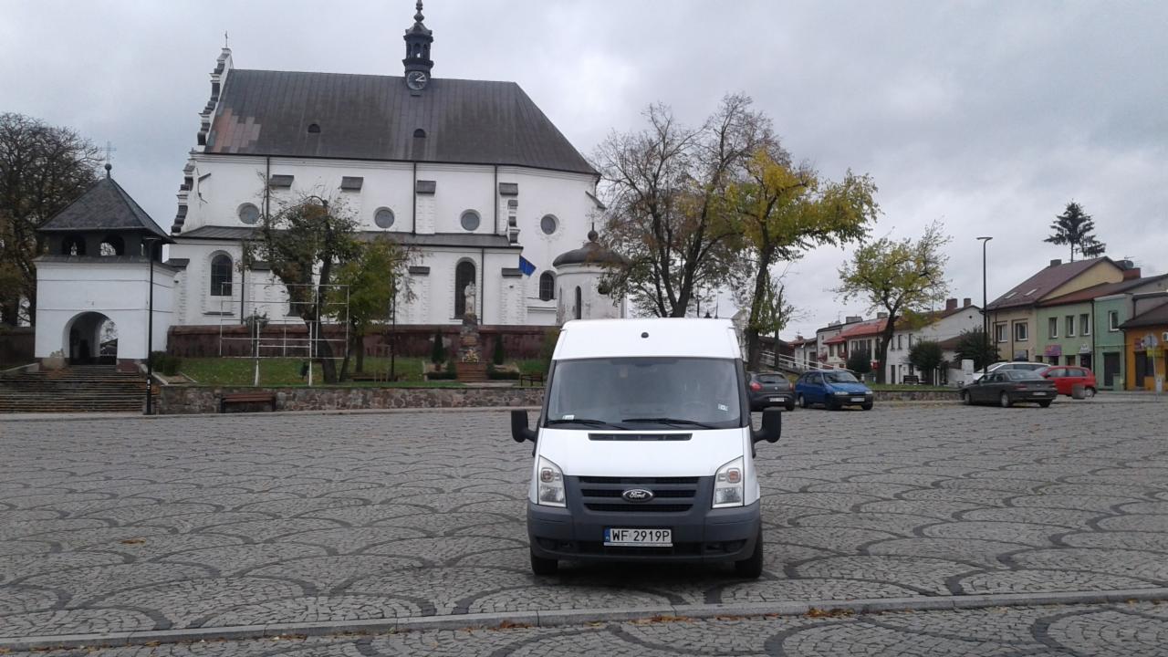 Biały bus Ford Transit parkuje na brukowanym placu miejskim przed białym kościołem z wieżą zegarową w pochmurny dzień.