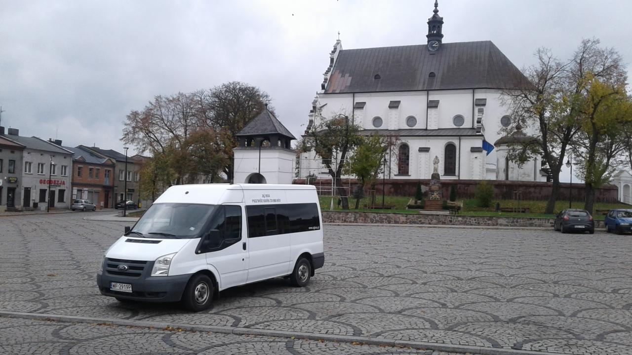 Biały bus transportowy Ford Transit na brukowanym placu, w tle biały kościół z wieżą i budynki miejskie w pochmurny dzień.