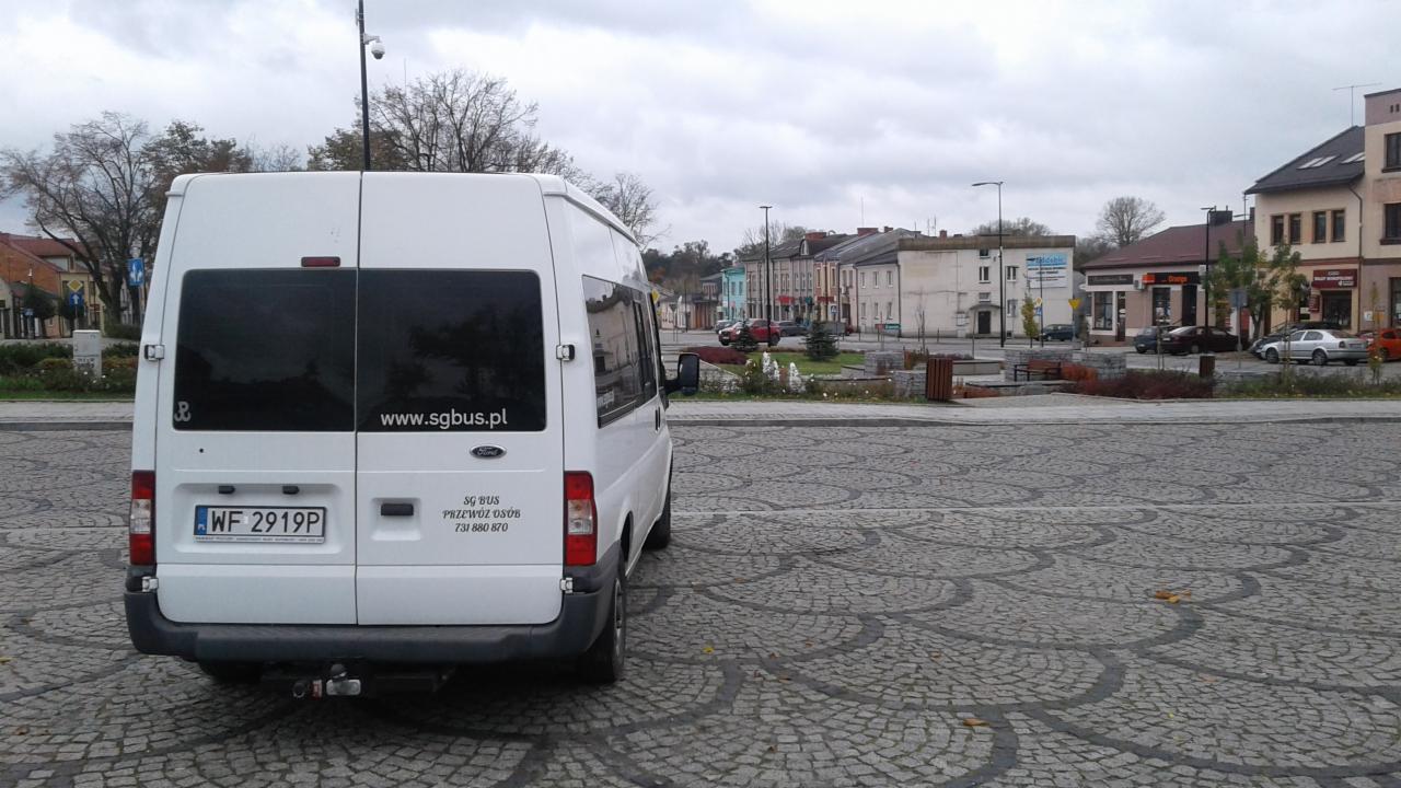Biały bus marki Ford z naklejką 'www.sgbus.pl' zaparkowany na brukowanym placu miejskim w pochmurny dzień.