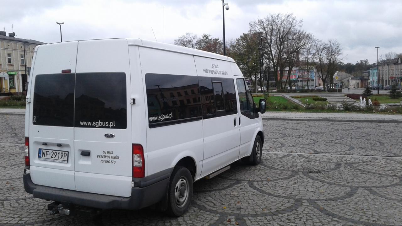 Biały bus firmy przewozowej z naklejoną stroną internetową www.sgbus.pl, zaparkowany na brukowanym placu miejskim z widoczną zielenią i budynkami w tle.