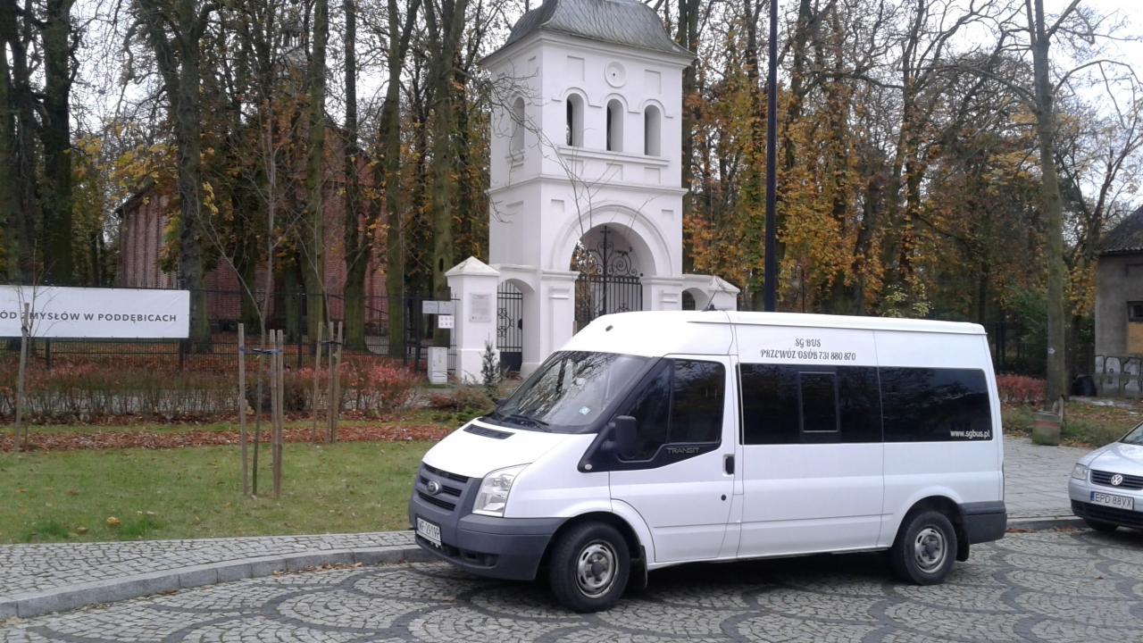 Biały bus Ford Transit z napisem 'Przewóz osób' i numerem telefonu, zaparkowany na brukowanej ulicy przed białym budynkiem z łukowym przejściem i metalową bramą, w tle drzewa z jesiennymi liśćmi.