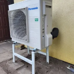 Zewnętrzna jednostka pompy ciepła Panasonic zamontowana na białym metalowym stelażu, z widocznym przyłączem rur, umieszczona na betonowym podłożu przy żółtej ścianie budynku.