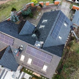 OZE EKOSYSTEM Sp. z o.o. - Widok z góry na dom z ciemną dachówką i panelami słonecznymi. Na dachu widoczna drabina i osoba. Obok domu zaparkowany biały samochód dostawczy.