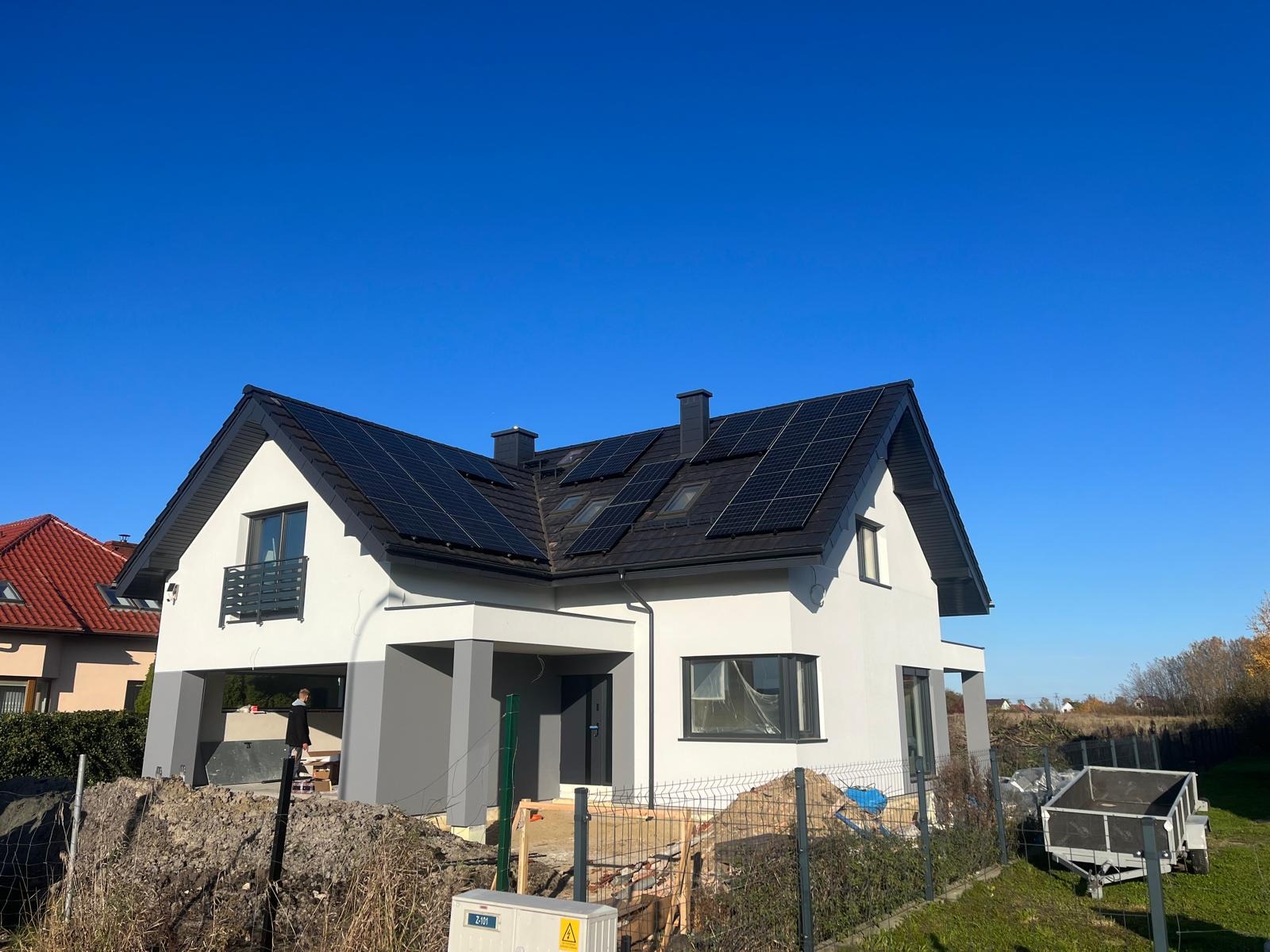 Biały dom z czarnym dachem i panelami fotowoltaicznymi w słoneczny dzień. Widoczny fragment działki i przyczepka.