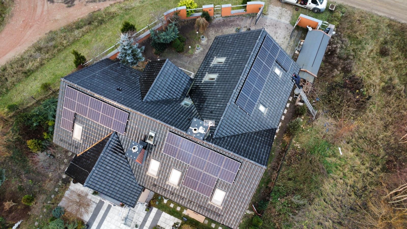 Widok z góry na dom z ciemną dachówką i panelami słonecznymi. Na dachu widoczna drabina i osoba. Obok domu zaparkowany biały samochód dostawczy.