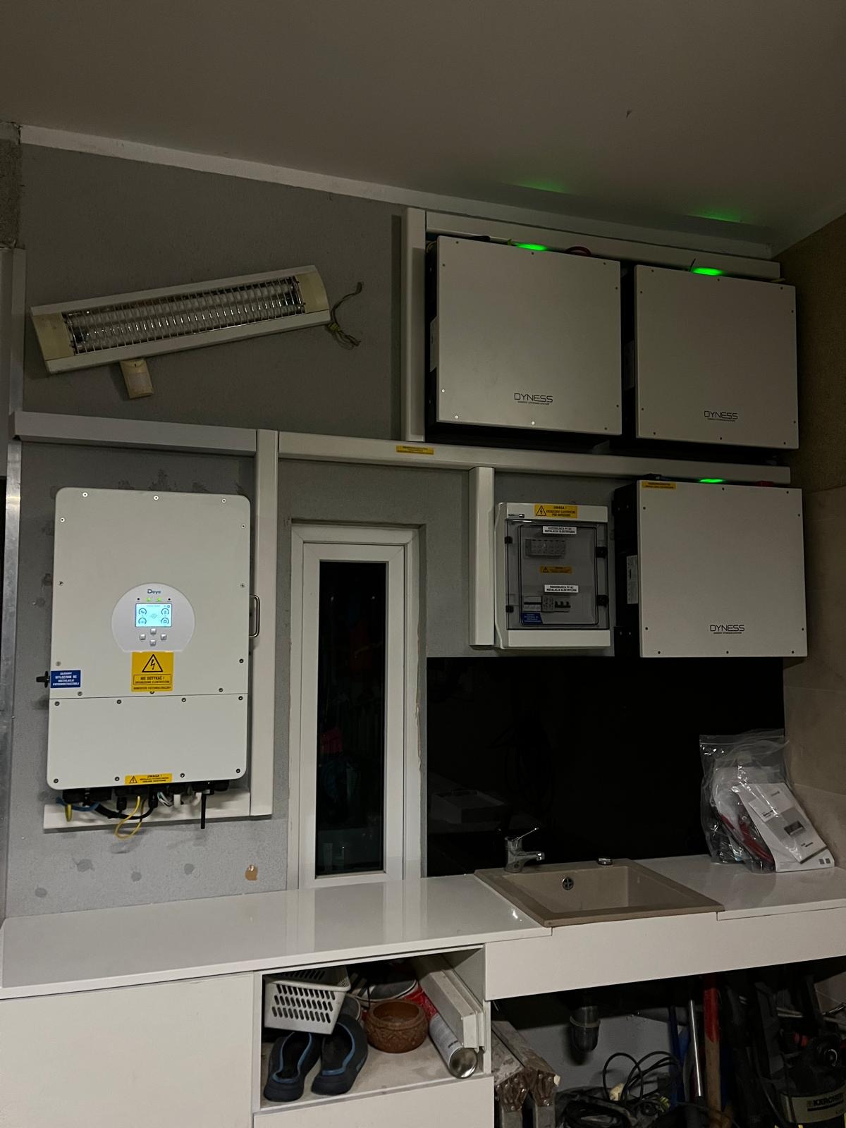 Instalacja fotowoltaiczna w pomieszczeniu: inwerter, akumulatory Dyness, rozdzielnica. Widoczny zlew i grzejnik. Nietypowe umiejscowienie.