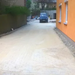 Świeżo ułożona, jasna kostka brukowa na podjeździe, widoczny samochód dostawczy i budynek z pomarańczową elewacją.