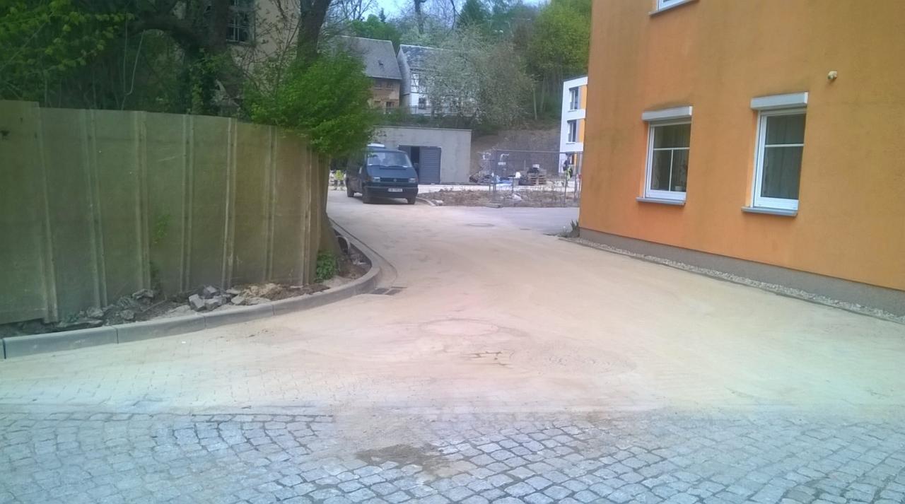 Przygotowanie podłoża pod kostkę brukową, widoczny piasek i fragment ułożonej kostki na obrzeżach, w tle budynek z pomarańczową elewacją i zaparkowany samochód dostawczy.