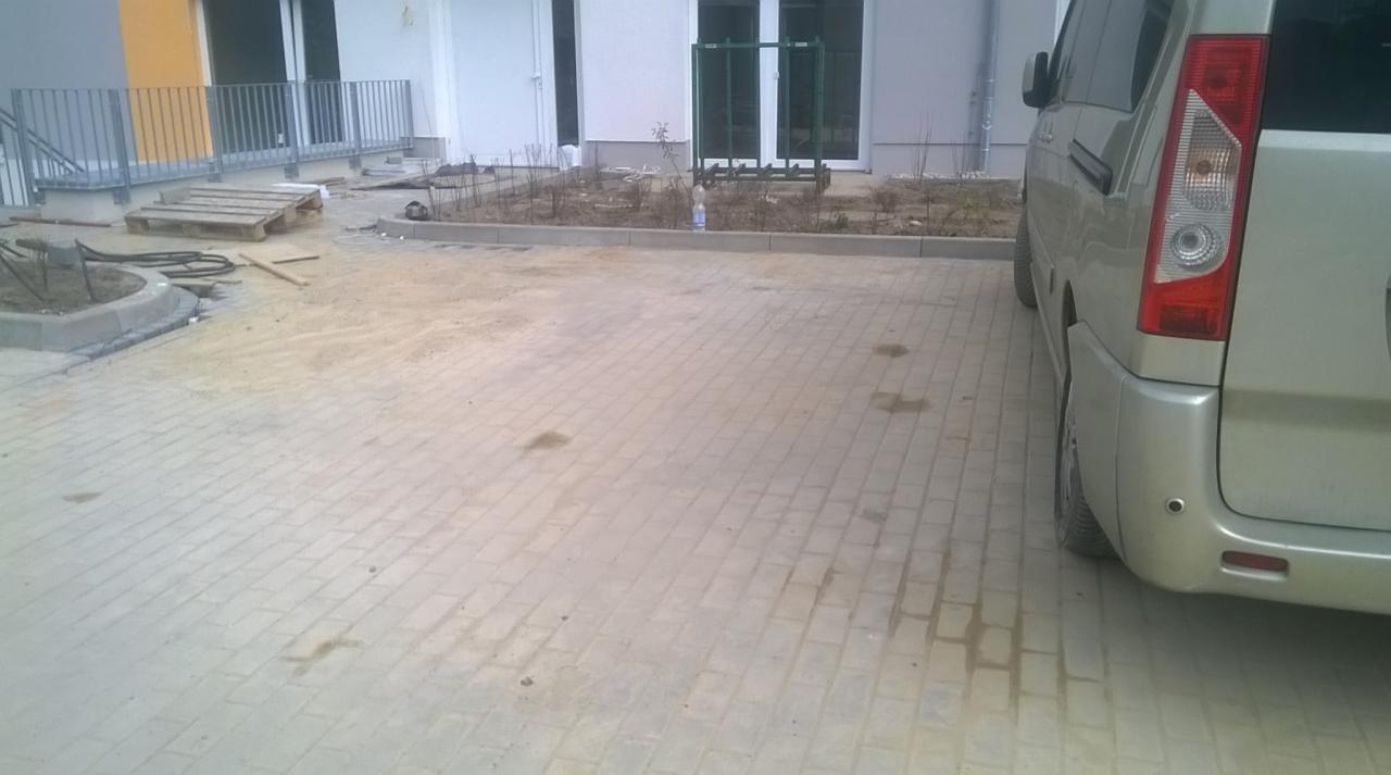 Ułożona kostka brukowa na podjeździe przed budynkiem mieszkalnym, widoczny fragment samochodu i paleta z materiałami budowlanymi.