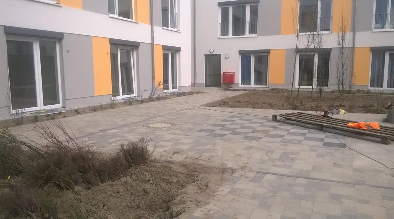 Dziedziniec nowego budynku z częściowo ułożoną kostką brukową w szachownicę; widoczne przygotowane podłoże, paleta z obrzeżami i odzież robocza.