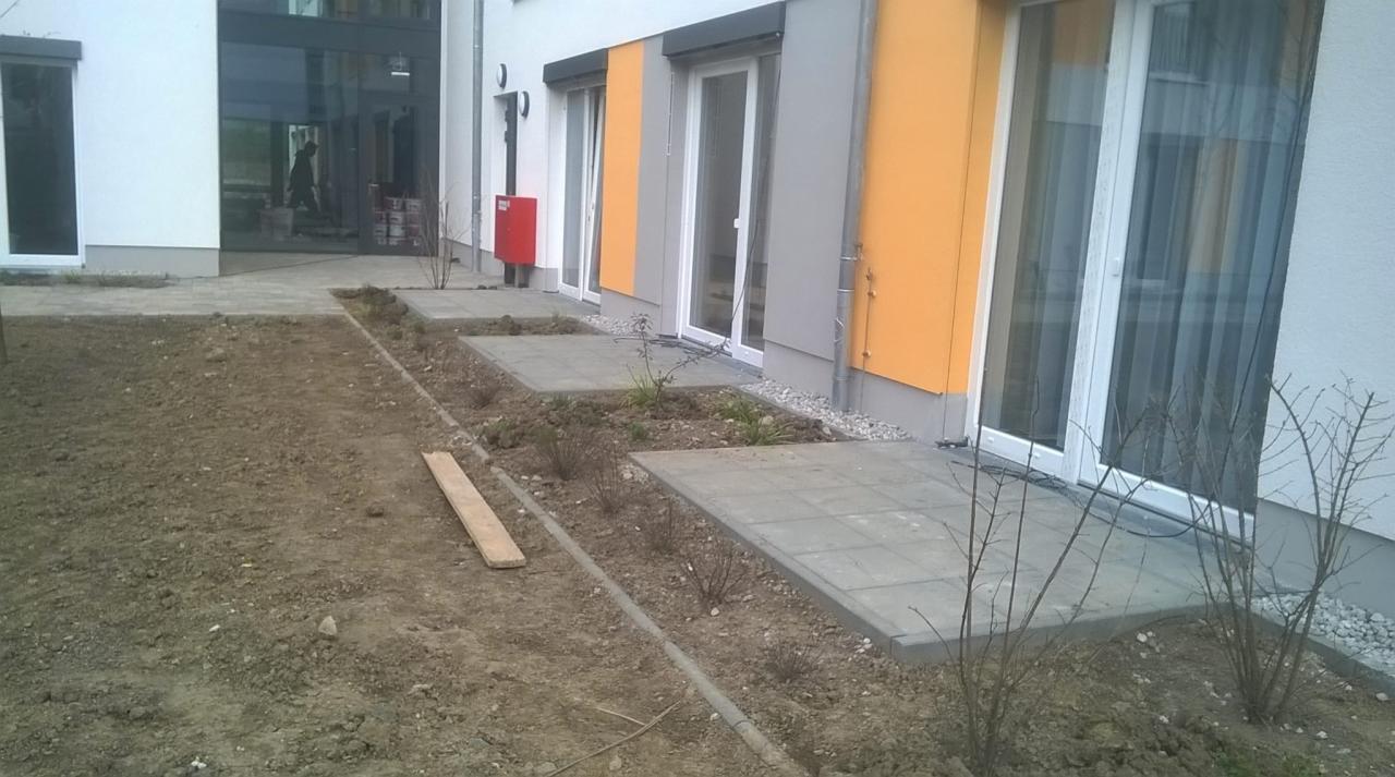 Wyłożone płytami chodnikowymi ścieżki prowadzące do wejść budynku, pomiędzy nimi rabaty z ziemią i sadzonkami, widoczne elementy elewacji w kolorach białym, szarym i pomarańczowym.