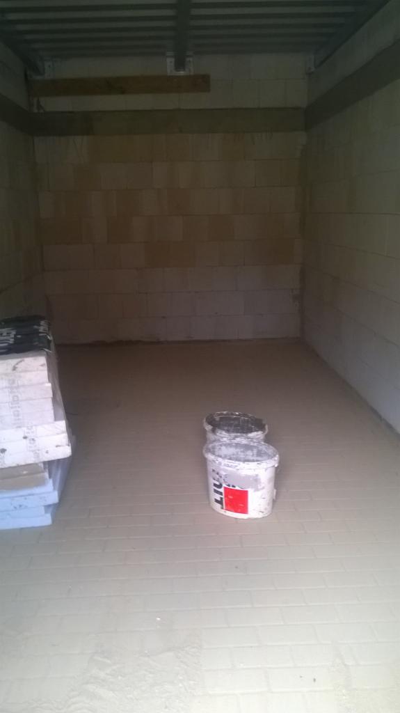 Wnętrze pomieszczenia z ułożoną jasną kostką brukową na podłodze, widoczne dwa wiadra z zaprawą i stos płyt styropianowych przy ścianie.