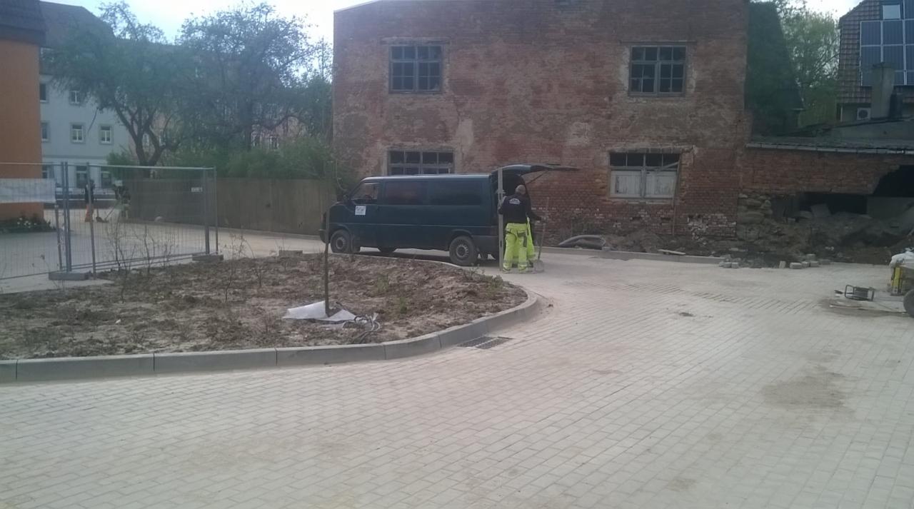 Pracownicy w odblaskowych ubraniach przy samochodzie dostawczym na tle starego ceglanego budynku, widoczna świeżo ułożona jasna kostka brukowa i przygotowany teren pod nasadzenia.