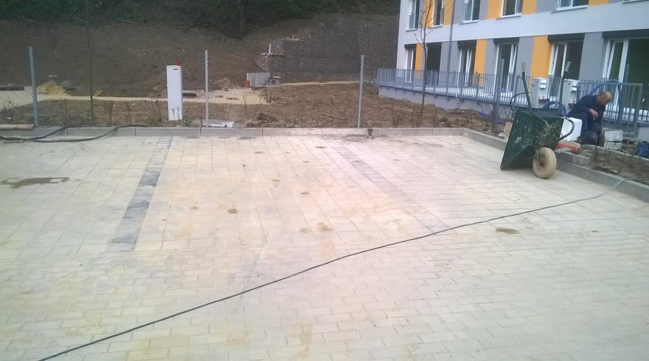 Układanie jasnej kostki brukowej z ciemniejszym pasem, widoczny pracownik przy krawężniku, taczka i nowy budynek w tle.