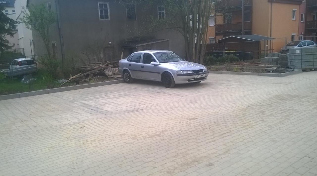 Nowo ułożona, jasnoszara kostka brukowa na podjeździe przed budynkiem mieszkalnym, z zaparkowanym srebrnym samochodem osobowym na środku. W tle widoczne stosy kostki brukowej i materiałów budowlanych.