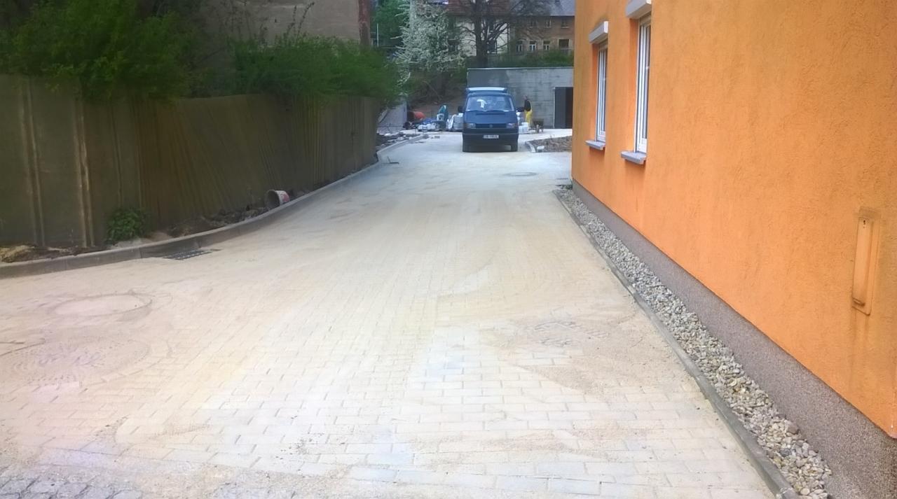 Ułożona kostka brukowa na podjeździe, widoczny samochód dostawczy i pomarańczowy budynek z dwoma oknami w tle.