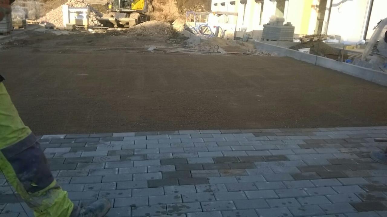 Fragment układanej szarej kostki brukowej na podsypce piaskowej, w tle plac budowy z koparką i stertą materiałów.