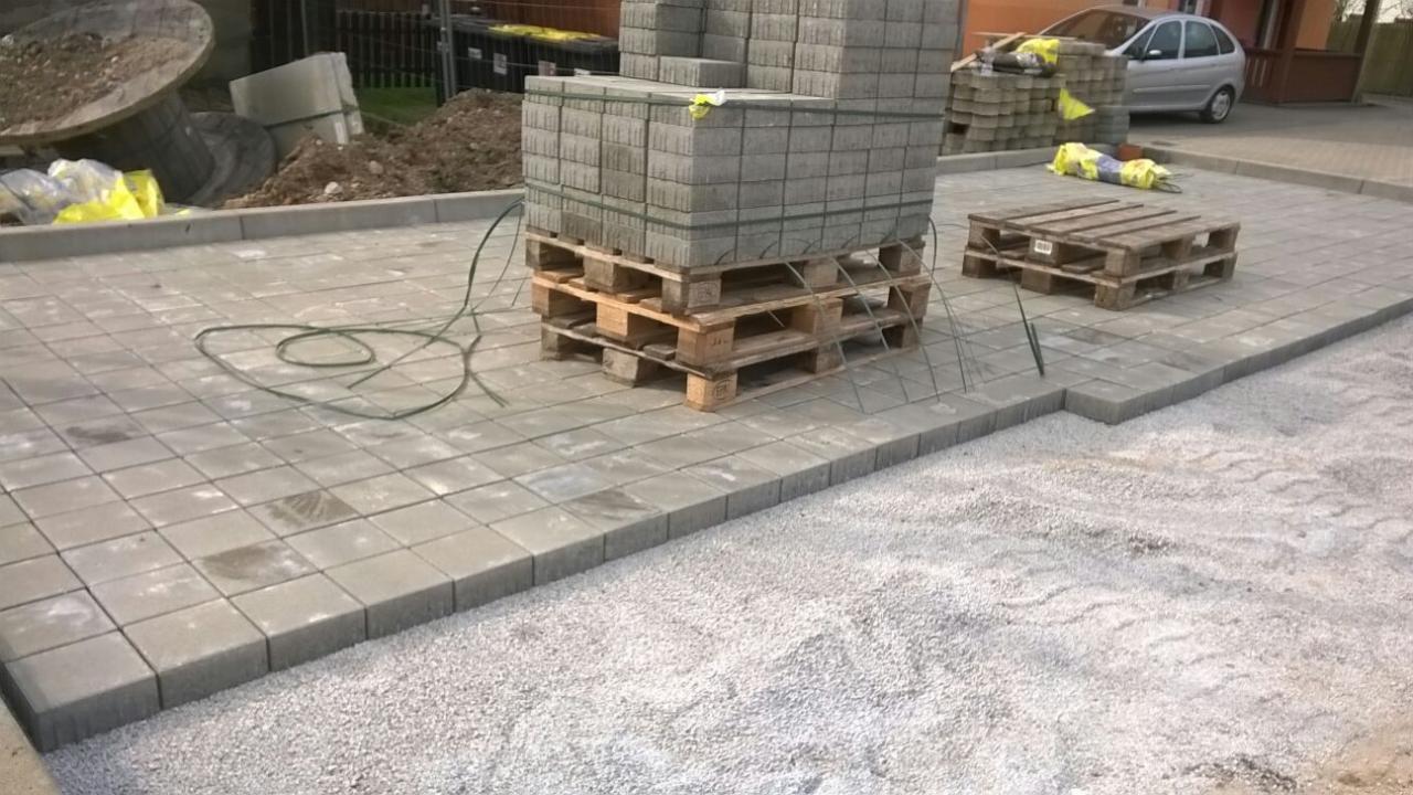 Fragment układanej nawierzchni z szarej kostki brukowej na podsypce z kruszywa; paleta z kostką w tle, widoczny fragment samochodu.