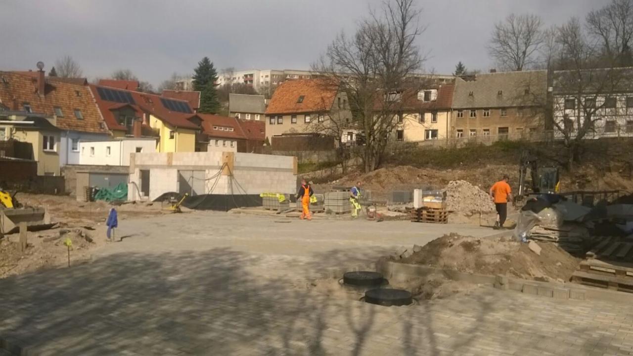 Obszar przygotowany pod budowę lub wyłożenie kostką brukową, z widocznymi pracownikami w odblaskowych ubraniach i stertami materiałów budowlanych na tle zabudowy miejskiej.