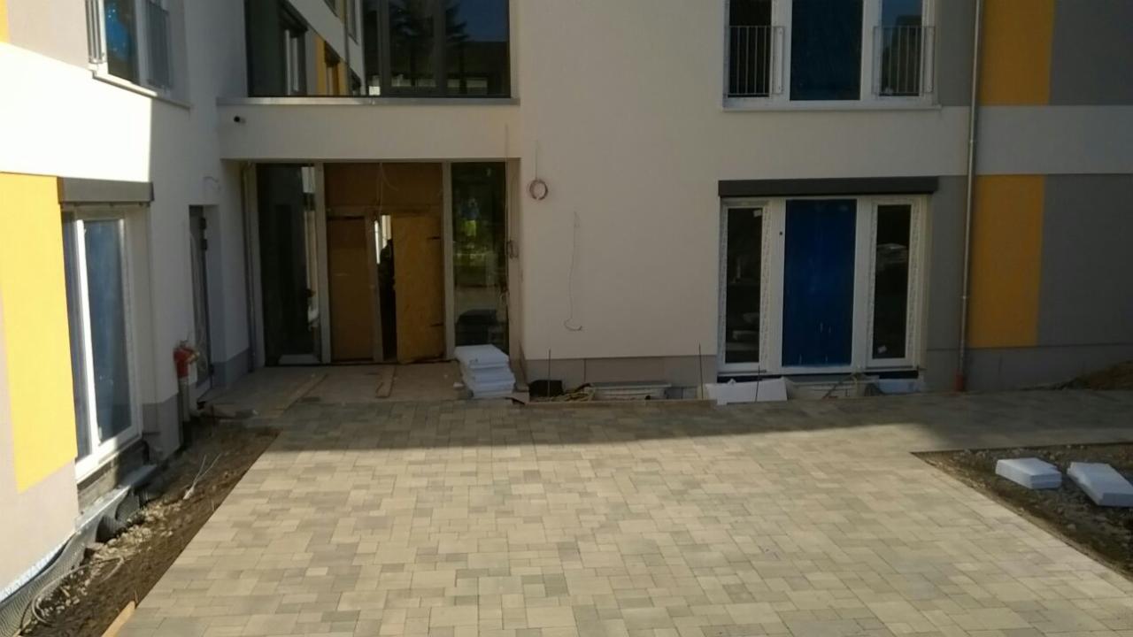 Nowo ułożona, beżowa kostka brukowa przed budynkiem mieszkalnym z żółto-szarymi elementami elewacji. Część kostki przy budynku jeszcze niezasypana ziemią.