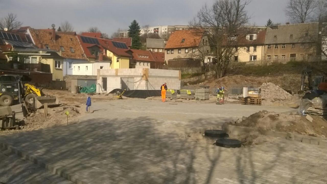 Rozległy plac z częściowo ułożoną szarą kostką brukową, w tle budynek w trakcie budowy, robotnicy w strojach roboczych i maszyny budowlane, widok z perspektywy lekko podwyższonej.