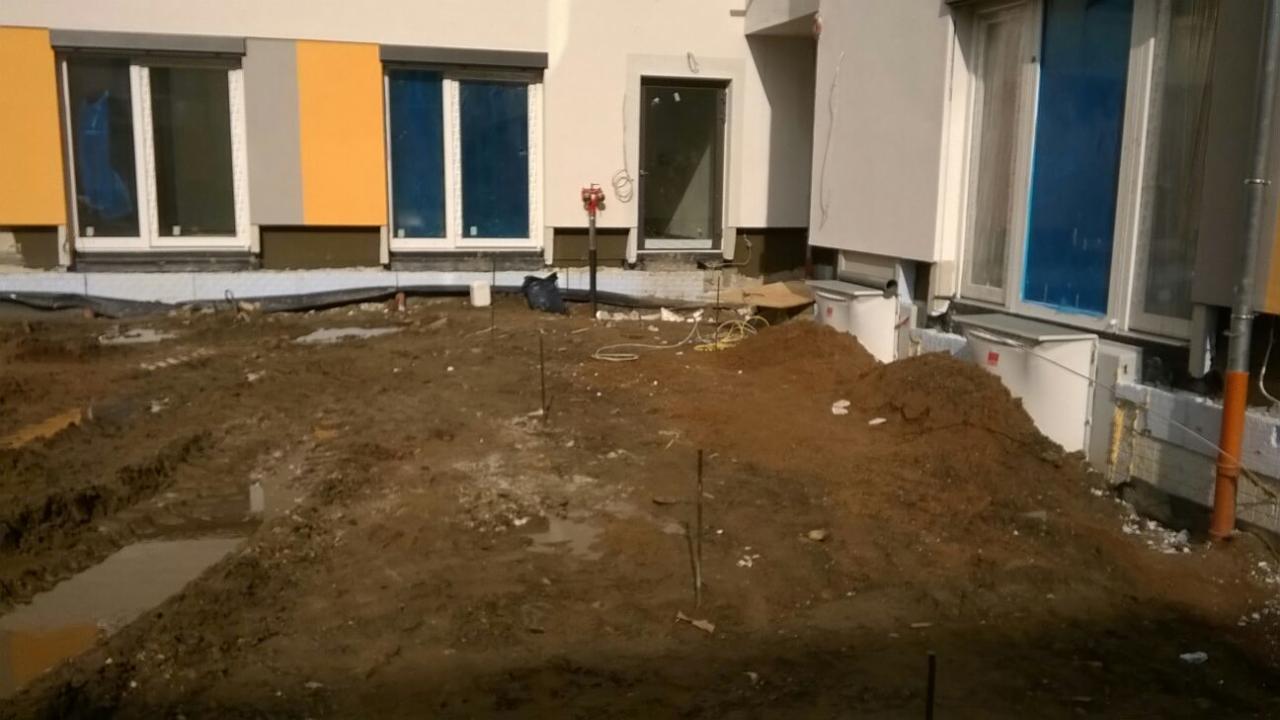 Przygotowanie terenu pod budowę: wykop z częściowo wylanym betonem, sterta ziemi, wystające pręty, widoczne okna budynku w tle.