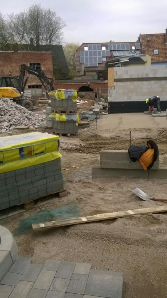 Prace brukarskie w Bydgoszczy w toku: ułożona część kostki brukowej, palety z kostką, wykop, koparka w tle, pracownik w żółtej kamizelce przycina kostkę.