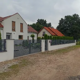 Nowoczesna, pozioma brama wjazdowa w kolorze grafitowym, zamontowana na białych słupkach, w tle dwa domy z czerwoną dachówką i zielone tuje, pochmurne niebo.