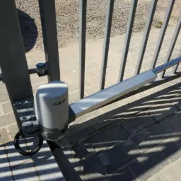 Szary napęd ramieniowy do bramy skrzydłowej zamontowany na metalowej bramie we Wronkach, widoczne detale mocowania i cień rzucany przez bramę na kostkę brukową.