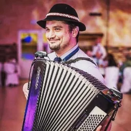 Uśmiechnięty mężczyzna w czarnym kapeluszu z dekoracją gra na akordeonie Weltmeister, w tle widoczni goście weselni i sala bankietowa.