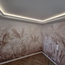 Pokój po remoncie: ściany z tapetą w motywy roślinne, listwa oświetleniowa pod sufitem i parkiet ułożony w jodełkę francuską. Subtelne, stonowane barwy.