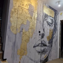 MJP Mateusz Wolny - Artystyczne malowanie ściany z motywem twarzy i złotymi akcentami. Widoczne niedoskonałości dodają charakteru. Oświetlenie podkreśla fakturę farby.