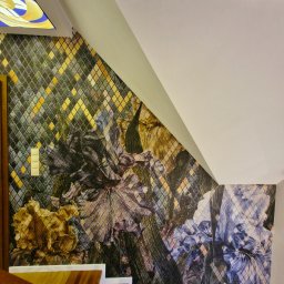 MJP Mateusz Wolny - Ściana w klatce schodowej pokryta tapetą w mozaikę z motywem kwiatowym, widoczny fragment schodów z drewnianą barierką i włącznik światła.